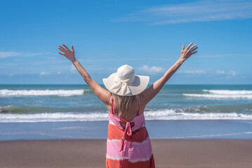 Back view of a mature woman raising her hands up while standing on the beach.