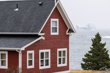 Fototapeta na wymiar A vibrant red colored two storey cottage style house with glass windows overlooking the blue ocean. In the distance, there's a large white glacier iceberg. The evening sky is pale blue and pink color.