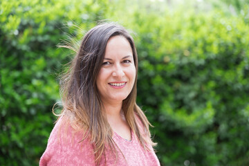 Outdoor portrait of a smiling 45 year old woman with windblown brown hair looking at the camera in the park on a sunny day. Confidence and tranquility of a mature woman. Happiness, lifestyle.