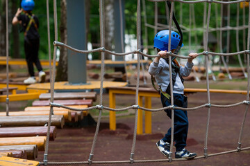 Summer. A small child climbs in a rope park on a rope bridge. A boy is having fun in an Adventure Park. A male baby on a climbing frame. Compliance with safety techniques.