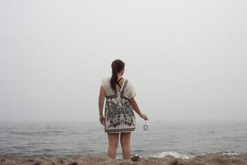 alone woman in a foggy beach with a lantern looking at the horizon
