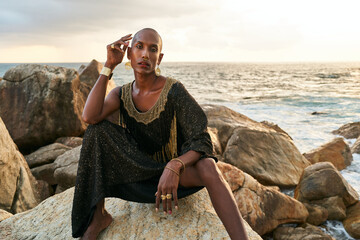 Non-binary ethnic fashion model in dress, brass jewelry sits on rocks by ocean . Trans sexual black person with rings, nose-ring, bracelets, earrings in posh clothes poses in tropical seaside location