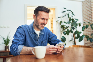 Fototapeta na wymiar Smiling adult man at home sitting at table using smart phone