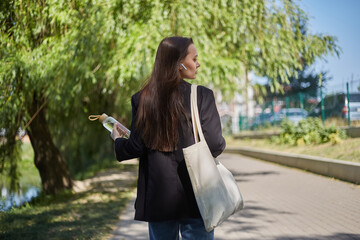 Beautiful young woman walking in the green park with a glass bottle of water in hand and cotton tote bag on a shoulder. Responsible and trendy female person on a walk