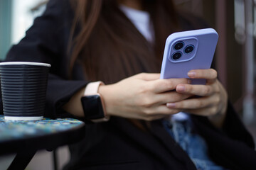 Young girl typing a message on a modern purple smart phone with triple camera. Unrecognizable female person drinking coffee and texting on a cellphone in a outdoor cafe