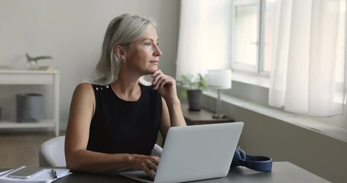 Busy mature freelancer woman working at home, using online application on laptop, typing, looking away with thoughtful face, thinking on creative text for writing articles