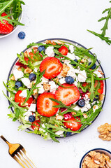 Fototapeta na wymiar Strawberry salad with arugula, lettuce, blueberries, feta cheese and walnuts, white table. Fresh useful dish for healthy eating