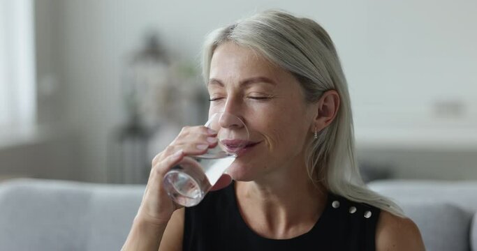 Peaceful positive mature woman drinking fresh clean pure water, holding transparent glass, keeping hydration balance for dry skin care, healthy metabolism, opening closed eyes, looking away, smiling