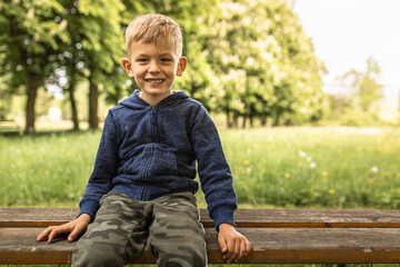 A smiling toddler blond boy sits on park bench, looking at camera.