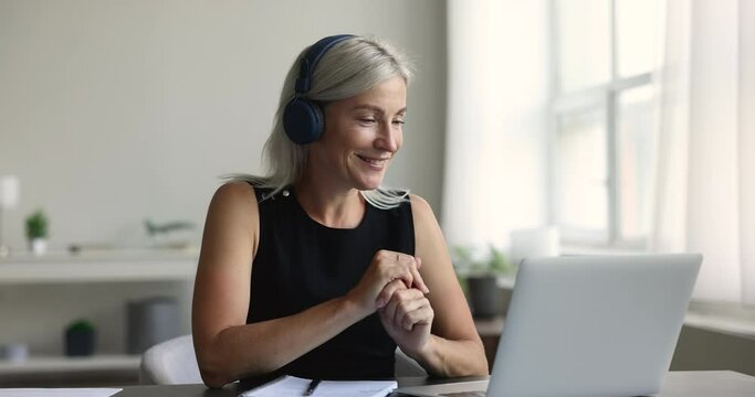 Serious senior woman in wireless headphones speaking on video chat call at laptop, working from home. Freelance manager, business lady talking to customer at computer, giving consultation