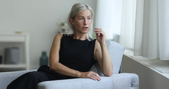 Thoughtful sad mature woman sitting in armchair at home, thinking over bad news, problem solving, feeling concerned, anxious, stressed, looking away, touching head, face, speaking