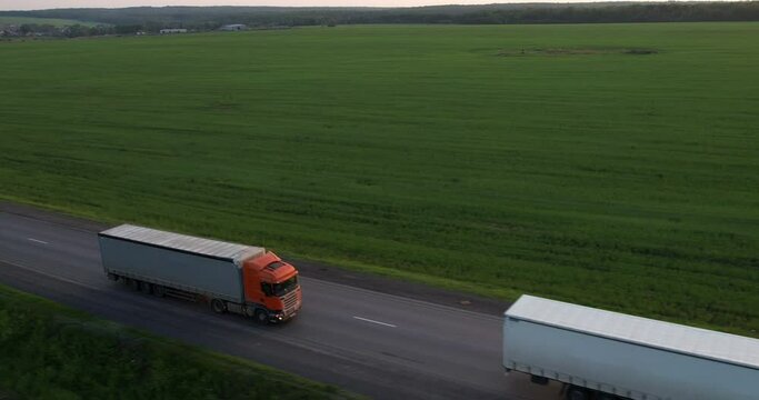 Trucks with a semi-trailer drive one after another along the highway, aerial shot. Freeway truck traffic at sunset in summer. Drone view. Cargo transportation. Green field