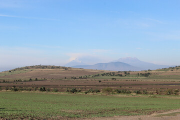 Fototapeta na wymiar Paisaje con campos, colina y volcanes de fondo con cielo azul. 