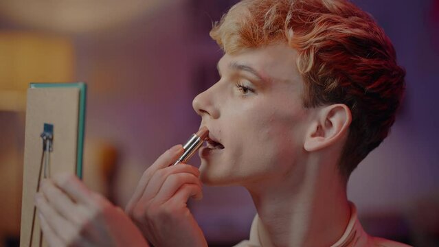 Close-up young non-binary person applying lipstick on lips, looking at his mirror reflection