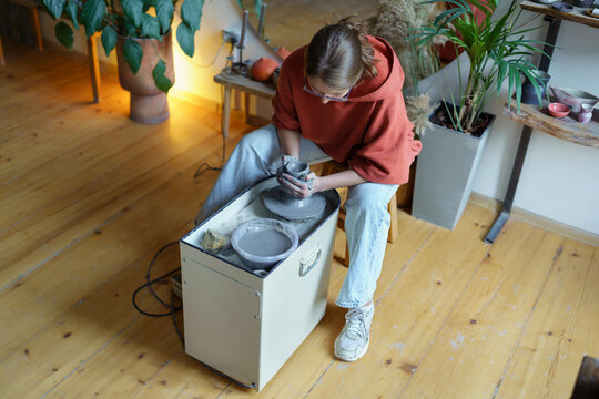 Focused businesswoman spends free time in ceramic workshop learning to make handmade dishes. Concentrated young female sits uses potter wheel getting rid of stress by making jars, pot, dish from clay