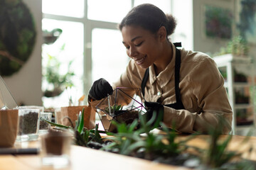 Black woman gardener transplanting succulents into glass vase creating florarium, arrangement green...