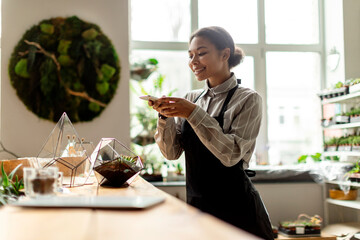 Young black woman gardener wearing apron taking pictures of plants on mobile phone while working in greenhouse, making modern florarium