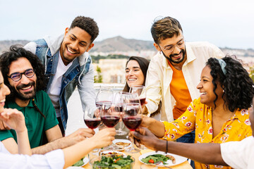 Group of young smiling people toast with red wine glasses at terrace party. Happy friends laughing and having fun enjoying lunch bbq celebration on rooftop in summer.