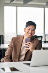 Young happy smiling professional Asian Japanese business man manager executive holding smartphone using mobile cell phone looking at camera working in office with technology devices, vertical.