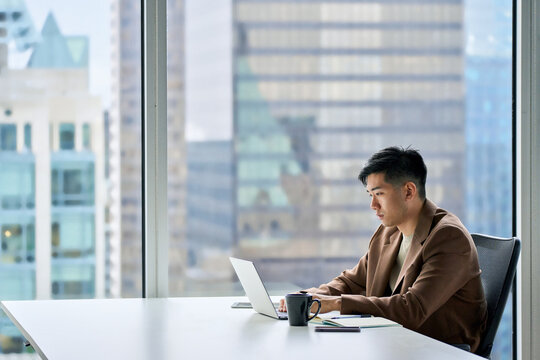 Young adult serious busy Asian business man manager wearing suit typing on laptop using computer working on digital project financial market data software management sitting at desk in office.