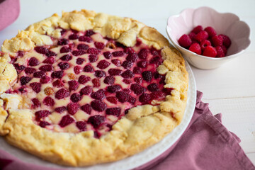 Homemade raspberry cake or galette on white wooden table over a pink cloth, with fresh berries on side, side view, close up.