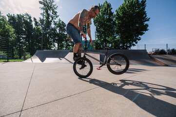 An oldish shirtless tattooed man is performing freestyle tricks on his bmx in a skate park.