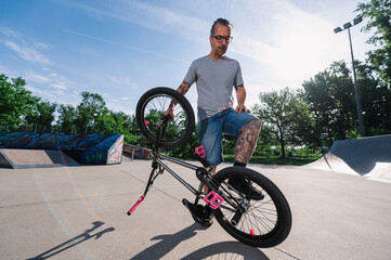 An urban active mature tattooed man is standing with his bmx bike in a skate park and practicing...