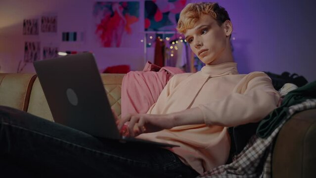 Relaxed charming guy with makeup using laptop at home with neon lights. LGBT. Gender fluidity
