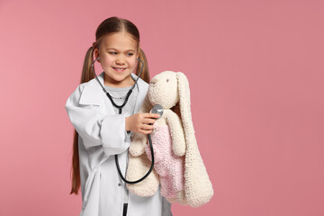 Little girl in medical uniform examining toy bunny with stethoscope on pink background. Space for...