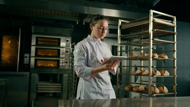 a woman baker in a uniform near the shelves with freshly baked bread holds a tablet in her hands checks bread bakery