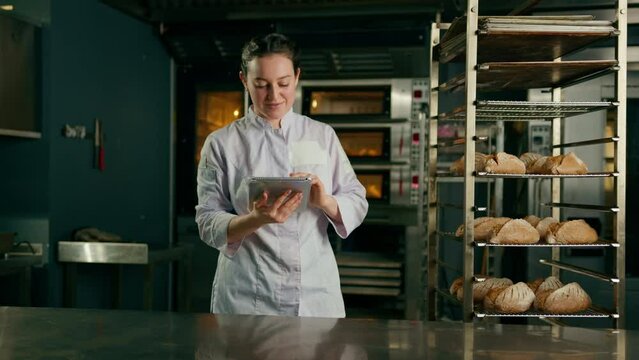 a woman baker in a uniform near the shelves with freshly baked bread holds a tablet in her hands checks bread bakery