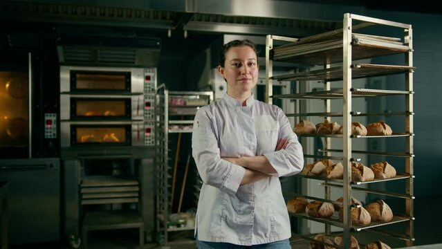 baker woman in uniform near shelves with freshly baked bread looking at camera and smiling