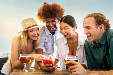 Young African American man and his happy multiracial friends looking at mobile phone in a beach bar while drinking beer.