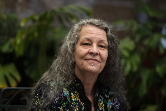 With a casual smile, a person relaxes in their garden. They have face piercings, which subtly allude to their self-described queer gender identity. Their silver hair shows themes of maturity and age.