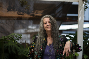 With one hand on a fence and a sun shade fabric in the background, a person relaxes in their backyard. They have flowing gray hair and a nose piercing.