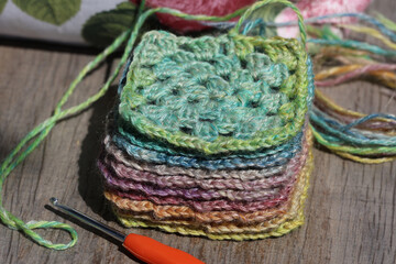 Closeup of a pile of crochet granny squares in mute rainbow colours with a crochet hook on a wooden...