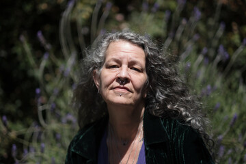 In an intense, sunlit portrait, a person poses next to a large lavender bush in their backyard. The sun makes their long, silver hair shine brightly.