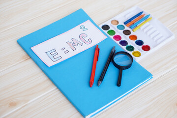 Paper card of formula E=MC2 on colorful books with pens, water color tray, crayons and magnifying...