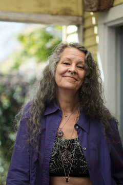 Smiling joyfully, an LGBTQ person poses in their backyard. They have a purple top, chunky jewelry, and nose and chin piercings. Their long silver hair is a testament to their maturity.