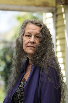 Looking over their shoulder, an older LGBTQ person looks seriously at the camera. They have long silver hair, face piercings, and chunky jewelry. They are at peace in their backyard.