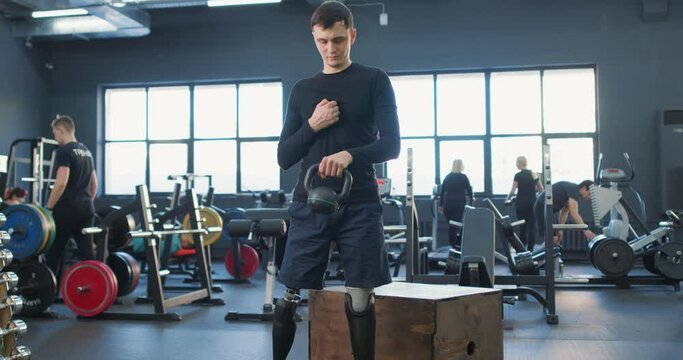 serious man with prosthetic leg standing in modern gym concentrated on doing exercises with kettlebell Slow motion. sportsman with artificial leg limb training with kettlebell in gym hobby interests
