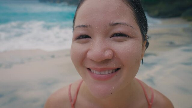 Close-up of Asian girl with sand on her face and wet hair looking at camera and tricky smiling against background of a tropical beach. Positive emotions happy Korean girl in swimsuit on vacation.