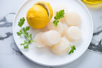 Fresh uncooked sea scallops with parsley and lemon on a white plate, horizontal shot on a white marble background, middle close-up