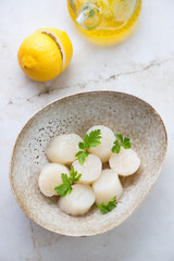 Bowl with raw fresh scallops, lemon and parsley on a light-beige marble background, vertical shot, high angle view