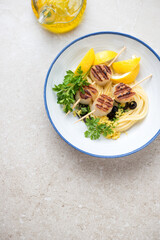 Grilled scallops on skewers with spaghetti, lemon and parsley in a white plate, above view on a beige stone background, vertical shot, copy space