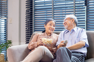 Senior asian couple is smiling while sitting on the sofa couch and having fun watching television...