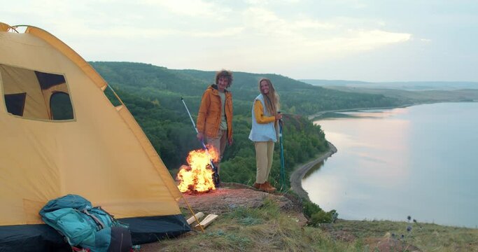 happy cheerful family couple is resting on the shore of lake with tent. Fashing camp with a fire in the forest. A man and woman travelling together, with trekking poles sticks. wonderful landscape