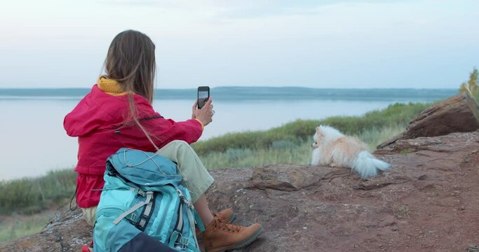 active female backpacker resting on stone, rock with her pet Spitz taking photo Slow motion. Young woman with her dog admiring mountains,lake on a sunny spring day Vacation holiday time concept
