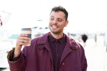 Young caucasian man holding a take away coffee at outdoors with happy expression