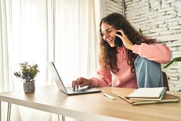 A young woman, focused and productive, working at her home desk with a laptop, multitasking while on a phone call.
